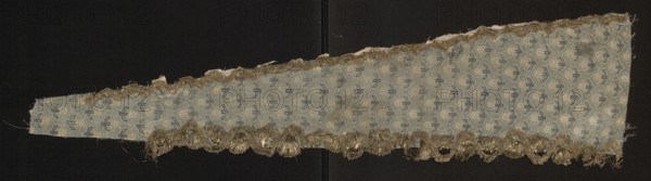 Fragment of a garment, fabrics with a preserved report, silk fabrics, floral patterns, fragments of a larger textile object. Design: small silver twigs form a net with meshes in the form of palmettes inside which white roses are placed; blue-silver stems. Dimensions reported: 3.7 x 2.7 unspecified units. Fabric density in 1 sq. cm: 31 white warps, 31 blue wefts. 17th century, Iran. Fabric height 76 cm, width 17.6 cm min. 4.3 cm., A long triangular strip of fabric laid against a dark background shows a repeated small floral or leaf pattern in muted blue and pale beige on a light bluish-gray ground, with a fringe of metallic gold threads along one long edge and a narrower metallic trim along the opposite long edge; the fabric edges are slightly frayed and the gold fringe appears tangled in places, and the overall colors visible are blue, beige, gray, gold, and dark black from the background.