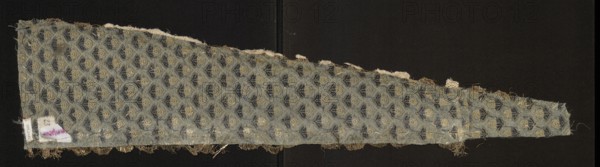 Fragment of a garment, fabrics with a preserved report, silk fabrics, floral patterns, fragments of a larger textile object. Design: small silver twigs form a net with meshes in the form of palmettes inside which white roses are placed; blue-silver stems. Report: 3.7 x 2.7. Fabric density in 1 square cm: 31 white warps, 31 blue wefts. 17th century, Iran, fabrics. Height 76 cm, width 17.6 cm min. 4.3 cm., A long, irregularly triangular fabric fragment laid flat against a dark background, showing a repeating small geometric motif that alternates between rounded and fan-like shapes; the fabric surface appears worn with some frayed edges and loose threads along the top and bottom borders, and a section of fringe remnants along one long edge. The visible colors include muted blue-gray as the dominant ground, darker navy or deep blue within the motifs, and a faded beige or tan accenting other parts of the pattern and the frayed fibers, with small patches of off-white where the material is mo...