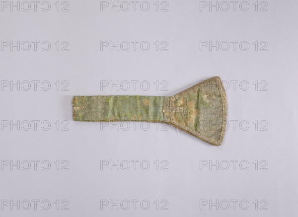 church paraments, liturgical vestments; plant ornaments; sacred art; churches and chapels furnishings; 1st half of the 18th century, France; liturgical vestments; length 68 cm; width 15 5.5 cm., An elongated fabric object laid flat on a light gray background, showing a long narrow strap section attached to a wider trapezoidal end; the fabric has a worn, textured appearance with visible stitching along the edges and a decorative woven pattern in shades of green, pale gold, muted beige, and hints of tan, with some frayed threads and slight discoloration.