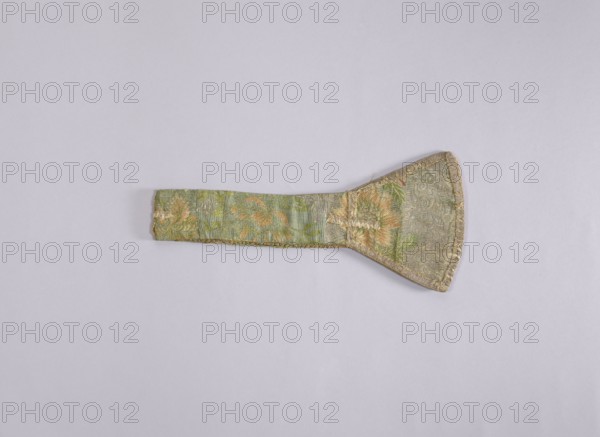 Manipulator, church paraments, liturgical vestments, plant ornaments, sacred art, churches and chapels furnishings, 1st half of the 18th century, France, liturgical vestments, length 68 cm, width 15 5.5 cm, A fabric object with a long narrow handle widening into a trapezoidal paddle shape lies flat on a pale lavender-gray background; the textile shows a woven floral pattern in muted green, gold, and peach tones with accents of darker green and a metallic sheen, and the edges are finished with a gold-colored trim.
