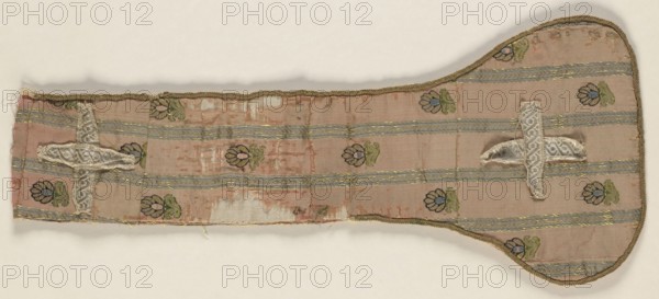 Fragment of a maniple made of crimson silk fabric with a striped and fine-thread motif. Salmon background, gold stripes and throws with black, green, and blue accents. The preserved fragment contains one ending pallette with slightly rounded edges trimmed throughout with a round wire. An isosceles cross made of silver braid is sewn onto the pallette. Identical fabric preserved in two fragments cut into stole pallettes. Fabric used for sewing zupans and kontusz lapels, probably from Grodno. Pattern motifs: small flower sprigs between gold stripes. Colors: pale red grosgrain background; trim: gold metal thread; brocades: pale blue, black, light green, gold, yellow. 2nd half of the 18th century, Poland. Liturgical vestment; length 43 cm, width 18 cm., A long, narrow fabric piece with a rounded wider end, primarily pink with a faded, worn appearance, showing a pattern of horizontal pale blue-gray stripes and small floral motifs in green, dark blue, and lighter blue. The fabric has metallic...