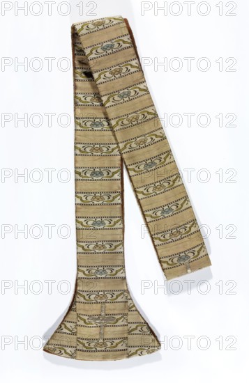 Stole, liturgical vestment. Stole originally part of a sash—still with transverse shelves, alternately plain and with a floral tendril motif. Ends palettes are sharply widened with slightly rounded lower edges, giving them a bell-shaped form. Crimson background with ornament in dark brown, creamy green, and dark blue. A fragment of a cross made of silver braid is sewn onto one end. Lining of yellow silk. 2nd half of the 18th century, Poland. Length 222 cm, width 21 cm., A long fabric band with a flared end laid on a white background, showing a repeating horizontal stripe pattern with decorative floral-like motifs; the fabric appears beige or tan as the base color with accents of muted green and pale blue in the motifs and darker brown outlining the pattern and edges, and a brownish inner lining visible along the folded edge.