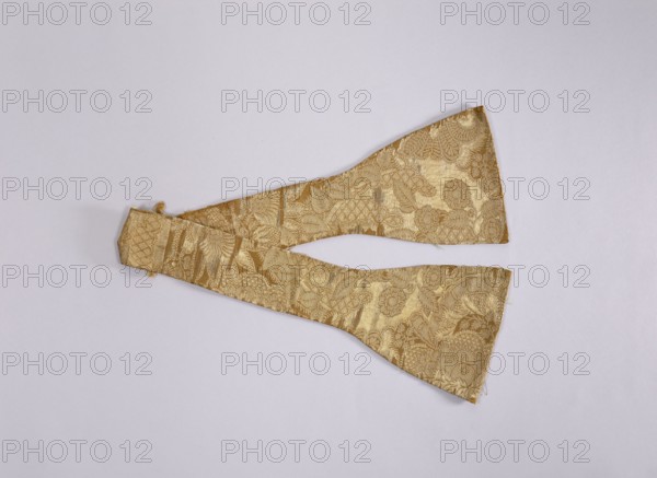 Manipulator, role of creator, church paraments; liturgical vestments; plant ornaments; sacred art; churches and chapels furnishings; 18th century, France; Lyon; liturgical vestments; length 90 cm; width 16 cm, A gold brocade fabric piece with a floral and leaf pattern lies flat on a pale gray background; the fabric has a metallic sheen and shows a few irregular triangular extensions joined at a narrower central section, with visible woven texture and slightly frayed edges, and the colors visible are gold and pale gray.