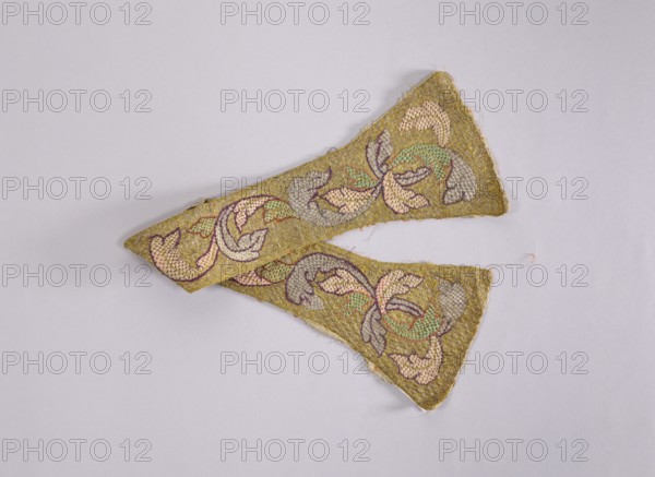 Maniple, liturgical vestment, early 18th century, Poland. Embroidered with a leafy twig motif in silver, green, pink, violet on a gold background. Unlined. Length 75 cm; width 16 cm. Cf. stole inventory number SZT 2480. Plant ornaments; sacred art; churches and chapels equipment., A pair of triangular fabric pieces with a textured, rough surface in a mustard yellow color, each decorated with a floral-like embroidered pattern in pastel pink, pale green, light gray, and a darker purple outline; the pieces overlap slightly and rest on a pale lavender-gray background, and there are a few loose threads visible at the edges.