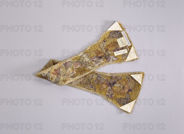 Maniple, gift; liturgical vestment, church parament. Embroidered with a leafy twig motif in silver, green, pink, violet on a gold background. Unlined. Early 18th century, Poland. Length 75 cm, width 16 cm., A triangular fabric strip with frayed edges lies on a pale gray background; the fabric shows a textured, worn surface with a mix of yellow, brown, purple, and muted red tones and areas of off-white and beige fibers, and there are a few small cream-colored triangular reinforcements attached at the corners.