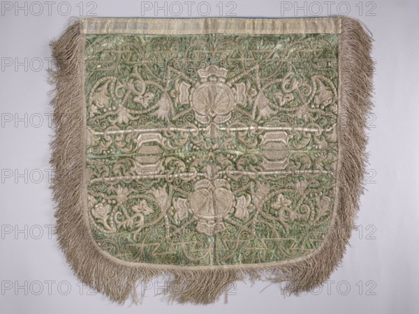 A hood from a church cope. The cope hood is made of pale green velvet. In the center, two embroidered palmettes run parallel to each other, surrounded by plant-braided motifs, with a finer ornament embroidered with a single thread closer to the edges. The hood is trimmed with silver braid: wider at the top, narrow around the remaining edges. The edges are fringed. 18th century, Poland. Liturgical vestment. Height 53 cm, width 52 cm., A rectangular textile with rounded bottom corners shown flat against a pale background, featuring a central area of green fabric with an intricate raised pattern in a light beige or cream color, surrounded by a fringe of long beige threads along three edges; the patterned area displays symmetrical swirling vegetal and geometric motifs in the cream thread against the green ground, with textured, slightly shiny fabric and visible stitching lines, and the top edge has a narrow band of lighter fabric in a pale beige or off-white tone.