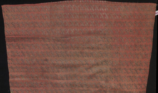 Fragment of fabric from a garment, Silk Factory Company, fabrics, silk, 2nd half 18th century. Decoration: small leafy twigs and two buds repeated in rows, alternating. Made on a wide loom over 78 cm; colors: crimson background with a rep weave; brocades in navy blue and ochre-silver. Pattern repeat: 4 x 2.5. Same fabric — PIECE 247. Place of origin: Grodno, Lithuania Grand Duchy of Lithuania; 1569–1795; Poland. Fabric, height 113 cm, width 39 cm., A rectangular piece of fabric with a muted reddish-pink background laid against a dark background, covered in a repeating diagonal vine-like pattern in darker grayish-blue and pale beige tones, showing subtle horizontal creases and slight variations in the red color with some areas appearing more faded or darker, and narrow black margins visible along the left and right edges.