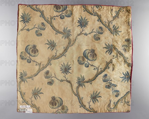 Velum;, revindication provenance; church vestments; embroidery; flowers, leaves, plant ornaments; sacred art; churches and chapels furnishings. Velum sewn from scarlet satin, embroidered with silk yarn and metal threads. In the center, a heart-shaped medallion features the letters IHS and a cross, with stylized floral sprigs on the sides. In the corners and along the sides are varied stylized flower bushes. The edges are trimmed with narrow gold braid. The lining is made of silk fabric brocaded with motifs of twigs and flowers and fruit growing from them on a light background. 18th century, Poland; liturgical vestment; height 49 cm, width 52 cm., A square piece of fabric with a pale beige background patterned with a repeating botanical design in shades of blue and muted green, showing curved branches with stylized flowers and round fruit-like forms; the fabric has a narrow red edging visible along two sides and a vertical seam where a narrower panel joins the main piece; the surface sh...