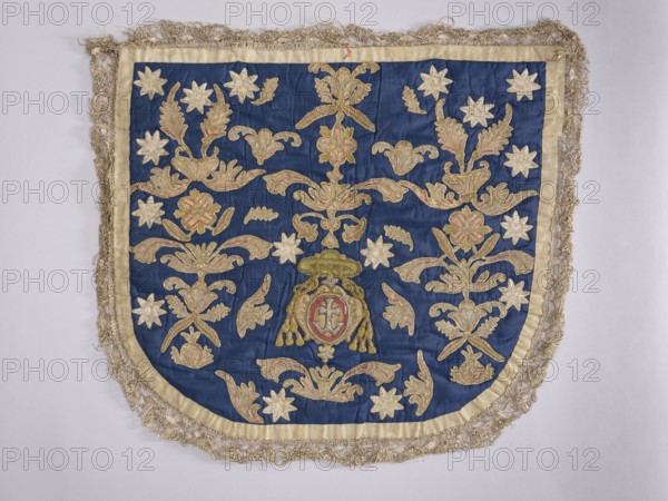A hood from a church cope. The hood of the liturgical cope is made of navy blue silk with appliquéd decoration of stylized flowers, leaves, and stars. In the lower center, beneath the bishop's hat, is a cartouche with the Odrowaz or Dolega coat of arms. Predominant colors are gold, silver, green, and brick red. The hood is trimmed with contemporary metal lace braid. The appliqué was transferred to a new fabric. 18th century, Poland. Fabrics. Height 58 cm, width 61 cm., An embroidered textile panel with a deep blue fabric background bordered by a pale gold trim and an outer fringe of metallic threads, featuring an array of ornamental floral and foliate motifs in shades of gold and muted pink set against the blue, a group of star-shaped floral forms in pale gold scattered across the surface, and a central decorative emblem with gold elements and touches of muted pink and red surrounded by symmetrical scrolling leaf and petal designs.