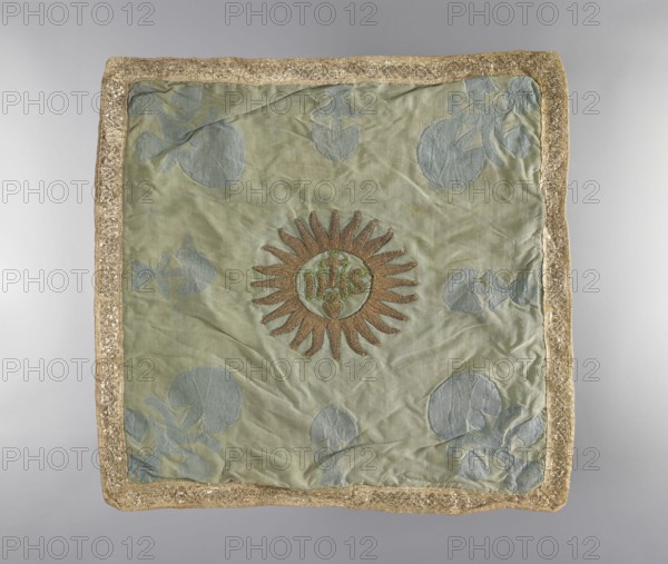 Velum., Velum made of blue silk fabric. In the center is an applied radiant medallion with the IHS monogram with a cross and a burning heart. In the corners and in the center of each side are traces of torn-out applied flower sprigs. Trimmed with wide silver braid. Linen lining. 18th century, Poland. Liturgical vestment, height 45 cm, width 46.5 cm., A square textile panel with a pale green background and a gold decorative border; the center features a sunburst motif with brownish-gold rays and a round inner medallion, and the fabric shows a subtle pattern of pale blue circular and leaf-like shapes scattered across the surface, with visible folds and slight wrinkles in the material.