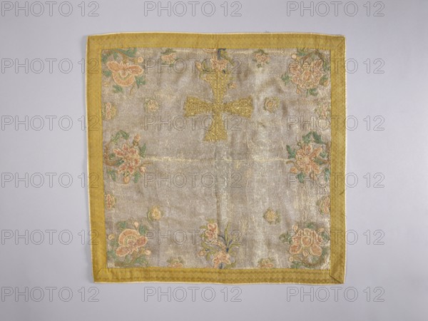 Velum, church vestment, floral motifs, flowers, leaves, plant ornaments, sacred art, churches and chapels furnishings. Velum made of silver lamé, embroidered in the corners and along the edges with colorful floral branch motifs in green, pink, salmon pink and beige. A gold equal-armed cross is applied at the center bottom. Edges trimmed with galloon; cotton lining. Early 18th century, Poland. Liturgical vestment, height 50 cm, width 50 cm., A square textile laid flat with a yellow border and a pale, slightly shiny central field. The central area shows a raised cross shape in a deeper yellow tone. Around the edges of the central field are floral embroidery motifs in shades of pink, peach, green, and a little blue. The background fabric appears silvery-beige with a subtle sheen, and the border has a textured yellow pattern. The overall colors visible are yellow, silvery-beige, pink, peach, green, and blue.