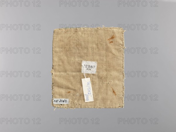 Palka, gift; church paraments; crosses; plant ornaments; sacred art; churches and chapels equipment; Bukowina; liturgical parament; height 15.5 cm; width 14 cm, A rectangular piece of rough beige fabric with frayed edges is centered against a plain light gray background; the fabric shows a few small brownish stains and some loose threads along its borders, and a small white tag is attached near the center. The visible colors are beige, light gray, white, brown, and a hint of blue on the tag.
