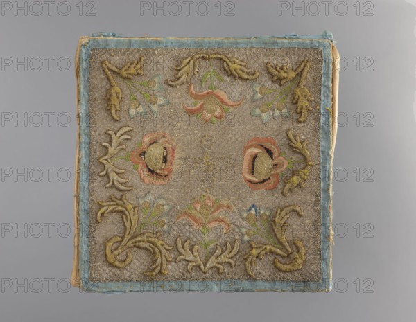 Embroidered pall; embroidery; church parament; liturgical parament; floral ornaments, flowers, leaves, plant ornaments; sacred art; churches and chapels furnishings. Pall embroidered with motifs of three types of floral branches placed in the center of each side and in the corners. Background set with silver metal thread sewn in a diagonal grid. Motifs embroidered with laid gold metal thread on a small backing and with satin stitch of colored silk yarn. In the center is a trace of a torn-out cross. Pall backed with cardboard, beneath which are visible fragments of old bills. 17th/18th century, Poland. Height 20 cm, width 20 cm., A square object with an embroidered surface framed by a worn light blue border, showing a textured beige background embroidered with floral motifs in pink, green, gold, pale blue, and hints of dark blue and black; the embroidery includes stylized flowers, leafy scrolls, and curling acanthus-like forms symmetrically arranged, with the fabric edges frayed in plac...