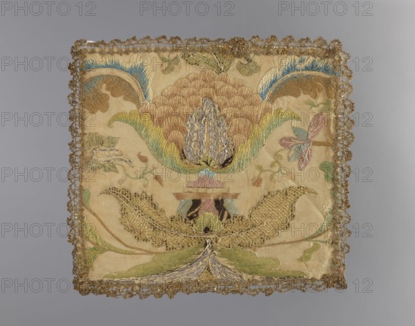Embroidered pall. Embroidery on silk fabric. In the center is a large palmette stylized flower on a base; large leaves grow from the palmette and base with branches bearing small flowers on the sides. Edges trimmed with narrow silver bobbin lace. Composition indicates it was made from a fragment of a larger embroidered fabric. 18th century, Poland. Liturgical parament. Height 22 cm; width 20 cm., A square embroidered textile with ornate scalloped metallic trim around the edges, showing a large stylized floral motif at its center rendered in shades of tan, beige, gold, green, blue, pink, and pale purple, with additional leafy and petal shapes in muted green and pink tones on a light beige background; the embroidery includes textured stitching and sections of metallic thread creating raised and filled areas, and a few smaller decorative elements and curling tendrils surround the central motif.