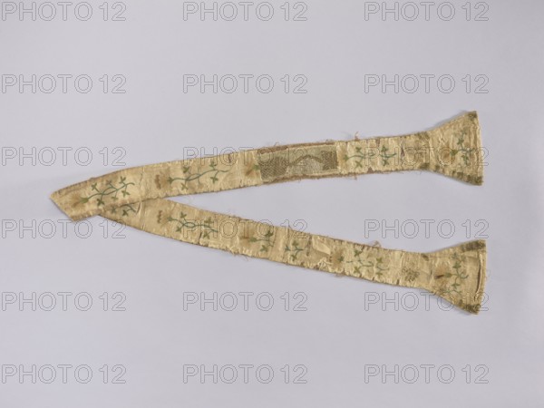 Stole, Church paraments; liturgical vestment made of silk, embroidered with carnation sprig motifs plant ornaments, flowers, leaves; sacred art; used in churches and chapels. Possibly related to a chasuble ca. 1750, Poland. Length 177 cm; width 14.5 6.5 cm., A long, narrow textile strip with widened ends lies against a pale background; the fabric is a light golden-beige with a slightly worn, frayed appearance and areas of darker gold sheen. A repeating vine-like motif in green runs along the length, accompanied by small floral shapes in muted gold and pale beige. The textile shows irregular patches of discoloration and slight tears revealing underlying threads, and the edges are uneven with loose fibers. The background is a soft, very light gray-lavender color.