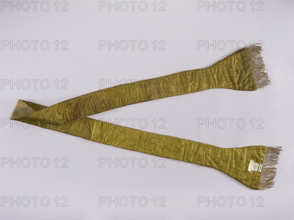 Stole, church paraments; flowers; liturgical vestments; plant ornaments; sacred art; churches and chapels furnishings. Stole embroidered with a leafy twig motif in silver, green, and two shades of pink. Gold fringe sewn to the ends of the paddles. 17th/18th century, Poland. Liturgical vestment; length 212 cm, width 19 cm., A long, narrow fabric band laid out in a loose V shape with fringed ends, showing a crinkled texture and uneven surface. The fabric appears mainly olive green with subtle variations in shade, and the fringe and some edges show a muted beige or tan tone. The background is a pale, light gray or off-white color.