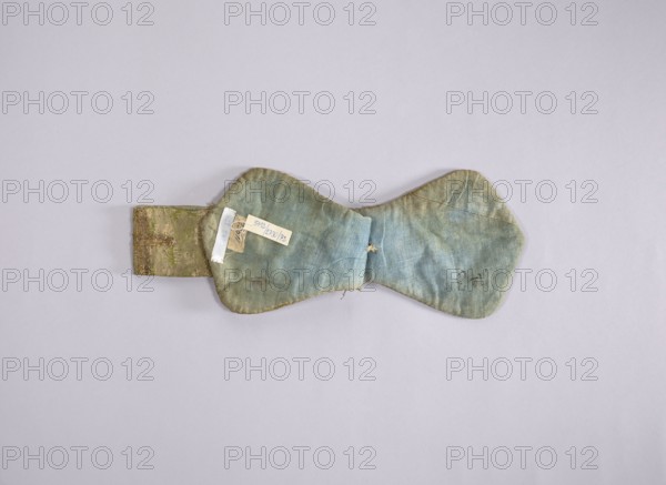 church paraments; flowers, leaves; liturgical vestments; plant ornaments; sacred art; churches and chapels furnishings; 18th century; France; liturgical vestments; length 81 cm; width 14 cm, A worn fabric object with a bow-tie shape lies flat on a pale grey background; the fabric shows faded blue and green tones with areas of beige and light brown discoloration, frayed edges, visible stitching, and small holes, and there is a short rectangular fabric tab attached on one side that has green and brown patterned fabric and a few small tags affixed to it.