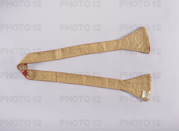 Stole, 18th century, Poland; liturgical vestment church paraments, embroidered sacred art; length 184 cm, width 18 cm. Description: discusses liturgical embroidery as sacred art—historical development of vestments, their form, symbolism, manufacturing techniques, significance of embroidery in Polish religious tradition, artistic and spiritual value; role of religious orders notably the Warsaw Capuchins in liturgical culture and care for vestments; analysis of theological content and aesthetic value of chasubles; descriptions of embroidery techniques illustrating the connection between craft, spirituality, and Church tradition., A long, narrow fabric strip arranged in a loose V shape on a pale background, the fabric appears mainly gold with a slightly shiny, textured surface and visible creases; the edges show a thin red trim and small areas of red backing visible at the ends; one end has a decorative applique in a pale metallic color, and the other end shows a few small light-colored t...