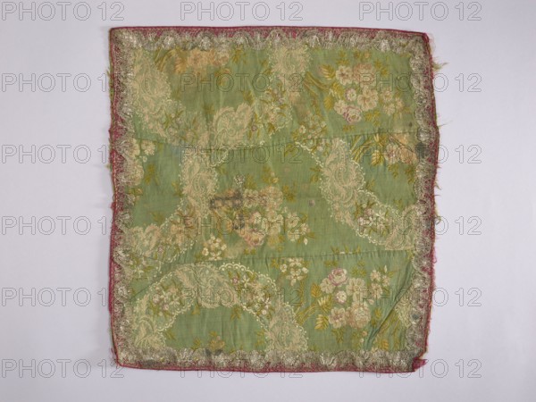 Velum; creator, church paraments; liturgical parament; flowers, leaves, plant ornaments; sacred art; churches and chapels furnishings; 2nd half of the 18th century; France; height 49 cm; width 45 cm; repossession provenance, A rectangular piece of fabric with a green background patterned with floral and paisley-like designs in pale cream, soft pink, and muted yellow, bordered by a narrow strip of faded red fabric and an ornate trim in metallic silver; the fabric shows areas of wear and small darkened spots, with a few creases and slight fraying along the edges, all set against a pale neutral background.