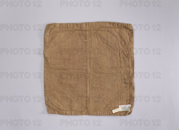 Velum; creator, provenance, church paraments; flowers, leaves, plant ornaments; sacred art; churches and chapels furnishings; 2nd half of the 18th century; place of origin, liturgical parament; height 42 cm; width 45 cm, A square piece of fabric laid flat on a pale gray background, the fabric is a light brown with visible creases and a slightly uneven texture, showing some faint darker spots and small scattered marks. The edges of the fabric are folded and stitched, and one corner has a small cluster of light-colored tags attached that contrast with the brown. The overall scene contains shades of brown, pale gray, and small areas of off-white.