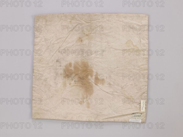 Vellum, vellum church parament, 18th–19th century, France; liturgical parament; height 44 cm, width 46 cm., A square piece of cloth lies flat against a pale gray background, showing a worn, off-white surface with visible creases and wrinkles; there are several brownish stains concentrated toward the center forming an irregular blotch, along with lighter discolorations and smudges scattered across the fabric; a few small darker specks and faint marks appear in different areas, and a small rectangular tag is attached near one corner; visible colors include off-white, beige, brown, gray, and a hint of cream.