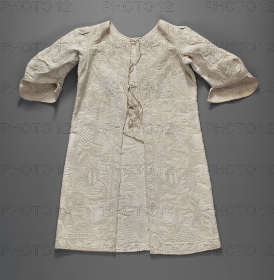 Baby's jacket, Repossession provenance. Clothing; children's clothing; baptismal garment. Baby's jacket made of weft-weave cotton fabric, decorated throughout with Saxon embroidery featuring motifs of stylized flowers, palmettes, and leaves twining on long branches. At the front, the main axis of the embroidery is marked by a row of heart-shaped palmettes. Among the stylized floral forms of embroidery are daffodils and carnations. Along the edges is a trail with a plant twig motif. Slit at the back and tied with four pairs of ribbons. Semi-circular neckline. Long, deeply set-in sleeves, finished with rolled cuffs, decorated with the Italian quilting technique. Unaltered. 1st half of the 18th century. Germany, Silesia. Fabrics. Length 47 cm; width 64 cm bottom., A long garment with sleeves laid flat against a smooth background, showing a textured, embroidered surface and a center opening with ties; the fabric appears off-white to cream with intricate raised floral and foliate patterns; ...