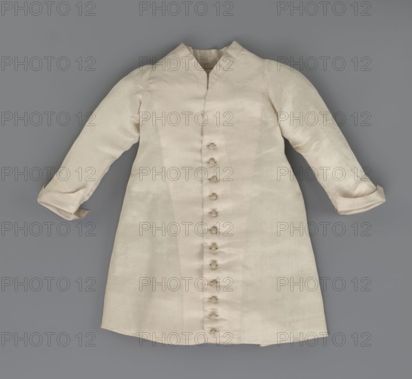 Children's dress made of cream satin. Long, fitted at the top, slightly flared at the bottom. Fastened at the front to the neck with 12 small, covered buttons. Low, stand-up collar. Long, set-in sleeves, finished with turn-up cuffs. Widened at the back, from waist level down, by gathered pleats. Unaltered. 2nd half of the 18th century, Poland. Garment; length 49 cm, width 100 cm bottom., A light-colored garment laid flat on a neutral gray background, showing long sleeves with turned cuffs and a V-shaped neckline, a central vertical row of small fabric-covered button loops, subtle vertical seam lines and paneling on the front, and a slightly flared skirt-like lower portion; visible colors include off-white, cream, beige, gray, and faint shadowed tones.