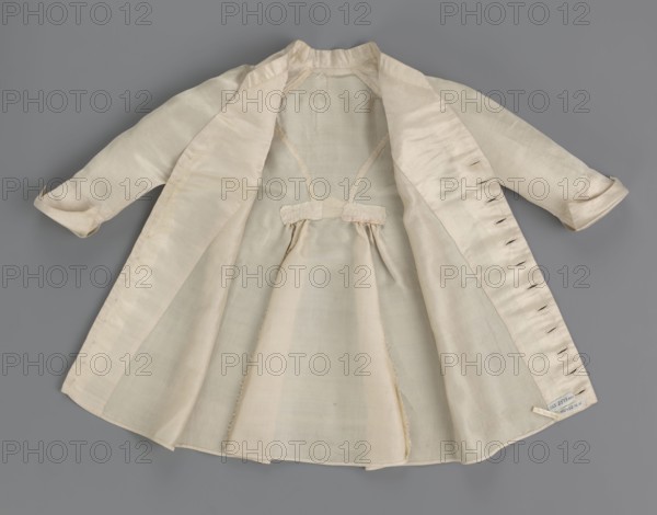 Children's dress made of cream satin. Long, fitted at the top, slightly flared at the bottom. Fastened at the front to the neck with 12 small, covered buttons. Low, stand-up collar. Long, set-in sleeves, finished with turn-up cuffs. Widened at the back, from waist level down, by gathered pleats. Unaltered. 2nd half of the 18th century, Poland. Garment; length 49 cm, width 100 cm bottom., A light-colored, open coat or dress laid flat on a gray background, showing the interior with a gathered skirt section and a small decorative tab at the waist; the garment has long sleeves with turned cuffs and visible seam lines and buttonholes along one front edge; the fabric appears smooth and slightly sheer in places, with subtle variations in shading; visible colors include cream, off-white, pale beige, and gray from the background.
