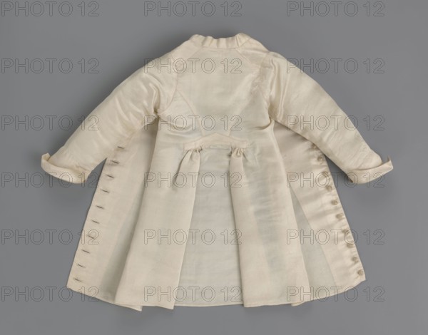 Children's dress made of cream satin. Long, fitted at the top, slightly flared at the bottom. Fastened at the front to the neck with 12 small, covered buttons. Low, stand-up collar. Long, set-in sleeves, finished with turn-up cuffs. Widened at the back, from waist level down, by gathered pleats. Unaltered. 2nd half of the 18th century, Poland. Garment, length 49 cm, width 100 cm bottom., A pale cream-colored long coat laid flat on a gray background, shown from the back with a fitted upper bodice and a flared skirt, featuring a gathered back waist detail and pleats in the skirt. The coat has long sleeves with turned cuffs, visible stitching and seams, and a row of small fabric-covered buttons along one side opening with corresponding buttonholes on the opposite side. The fabric appears smooth and slightly lustrous with subtle variations in shading. The background is a uniform medium gray.