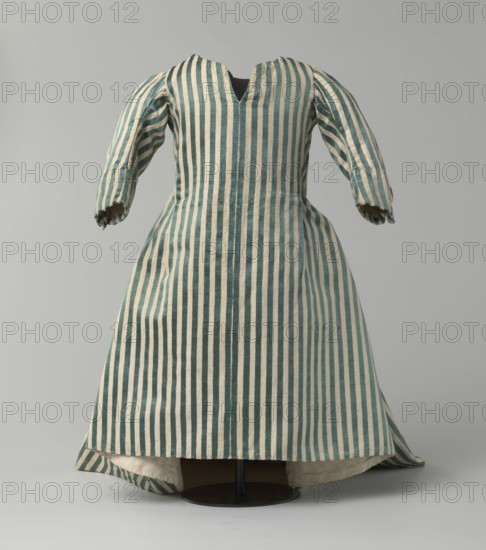Children's dress, 18th/19th century, Silesia. Made of satin with green and cream stripes. Fitted at the top, flared at the bottom, with a semicircular train extending at the back. Bodice slit at the back and laced. Neckline semicircular with a sharp V at center front. Long sleeves cut in an arc at elbow height, finished with small close-fitting cuffs fastened with covered buttons. Lining of white linen canvas. Unaltered. Length 49 cm., A garment displayed on a stand with a fitted bodice and long sleeves, made from vertically striped fabric in teal and off-white, with a slightly flared skirt and a small central opening at the neckline; the sleeves show the striped pattern running lengthwise and the skirt hangs to the base of the stand revealing an off-white inner lining; the background is a neutral light gray and the stand is a dark circular base.