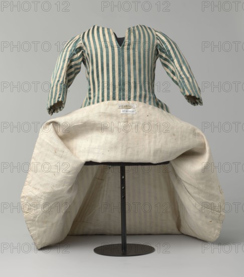 Children's dress, 18th/19th century, Silesia. Children's dress made of satin with green and cream stripes. Fitted at the top, flared at the bottom, extended at the back by a semicircular train. The bodice is slit at the back and laced. The neckline is semicircular with a sharp V-neck at the center front. Long sleeves, cut in an arc at elbow height, finished with small close-fitting cuffs fastened with covered buttons. Lining made of white linen canvas. Unaltered. Length 49 cm. Classification: clothing, children's clothing; fashion; provenance: repossession., A striped garment with vertical teal and cream stripes on the bodice and sleeves sits on a stand, showing puffy sleeves and a V-shaped neckline; a large cream-colored skirt or underskirt with a textured woven surface is folded outward at the waist, revealing the striped interior lining and a darker support structure beneath; the visible colors are teal, cream, off-white, dark gray, and black, and the background is a smooth light gr...