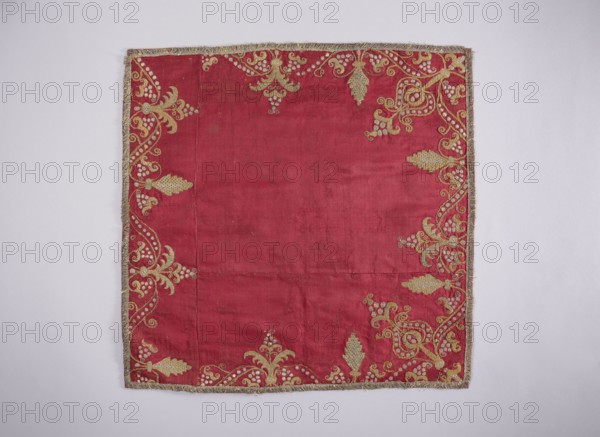 Velum of dark red satin embroidered around the edges with gold and silver thread in motifs of stylized flowers, trees, and palmettes. Edges trimmed with a short silver fringe. Lining of dark red silk moire. Early 18th century, Poland. Liturgical vestment, height 49 cm, width 49 cm., A square piece of fabric displayed flat against a pale background, the fabric is primarily deep red with a border of metallic gold fringe; the edges of the red area are decorated with an ornate gold embroidered pattern featuring flowing vine-like shapes, stylized leaves, and clusters of small round motifs, concentrated along the left, right, top, and bottom margins while the central area remains plain; the gold embroidery contrasts with the red ground and the fringe has a slightly tarnished metallic tone; the background around the fabric is light gray to off-white.