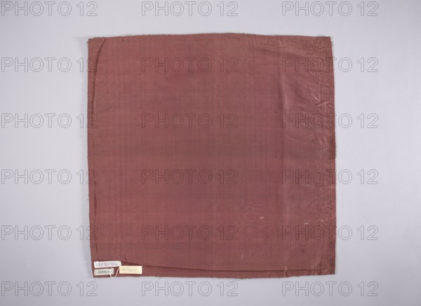 Velum of dark red satin embroidered around the edges with gold and silver thread in motifs of stylized flowers, trees, and palmettes; edges trimmed with a short silver fringe; lining of dark red silk moire. Early 18th century, Poland. Liturgical vestment; height 49 cm, width 49 cm. Makers/ Subjects: church vestments, flowers, leaves, plant ornaments, sacred art; intended for churches and chapels furnishings. Repossession, A square piece of fabric hangs against a light gray background, showing a muted reddish-brown color with subtle variations in tone and faint woven texture; the fabric has slightly uneven edges, a few small wrinkles and creases, and a narrow hem along the bottom edge, with a small lighter-colored tag cluster attached near the lower left corner.