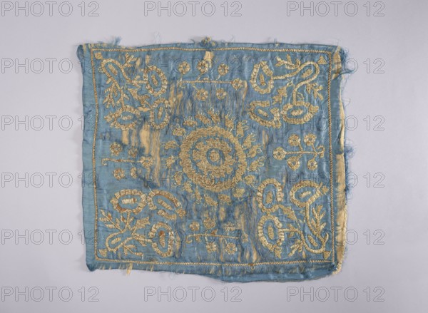Embroidered vellum, church paraments; flowers, leaves, plant ornaments; sacred art; churches and chapels furnishings. Velum made of blue silk satin, embroidered with silver thread. In the center, a rosette consisting of two ovals. In the corners, large, stylized, schematically treated bouquets of flowers, between which very simplified, smaller flower trees. For reinforcement, embroidered on beige woolen fabric. Lining detached. 2nd half of the 17th century, Poland; liturgical parament; height 52 cm, width 44 cm., A square piece of fabric with frayed edges laid flat on a pale background, predominantly light blue with metallic gold embroidery forming a central circular motif and repeating floral and looping decorative patterns toward the corners and edges; the blue cloth shows areas of wear and slight discoloration where the gold thread is denser, and the embroidery thread has a textured, raised appearance contrasting with the smoother fabric.