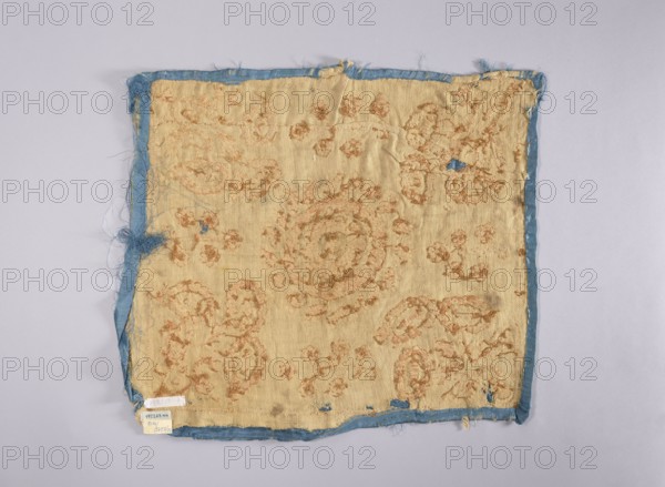 Embroidered vellum, Church paraments;, repossession Glogów. Flowers, leaves, plant ornaments; sacred art; churches and chapels furnishings. Velum made of blue silk satin, embroidered with silver thread. In the center, a rosette consisting of two ovals. In the corners, large, stylized, schematically treated bouquets of flowers, between which very simplified, smaller flower trees. For reinforcement, embroidered on beige woolen fabric. Lining detached. 2nd half of the 17th century, Poland. Liturgical parament. Height 52 cm, width 44 cm., A worn square piece of fabric lies flat against a light gray background, showing an overall faded beige surface with textured, raised patterns in a pale brown or tan color and a narrow frayed border in faded blue; the fabric has several small tears and areas where blue backing shows through, with loose threads and frayed edges visible around the perimeter and a few darker smudges on the beige surface.
