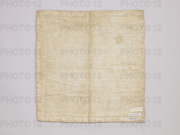 Vellum, vellum church parament with floral ornaments, satin vellum with floral ornament, late 19th century, Russia, Germany, liturgical parament, height 43 cm, width 42 cm, A rectangular piece of fabric lies flat against a neutral background, showing a faded, off-white to beige color with visible creases and folds across its surface; the fabric has slightly darker beige stitching along the edges and a few small irregular discoloration spots and a larger, irregular stain toward the upper right area; there is a group of small pale labels attached near the lower right corner, and the background appears light gray.