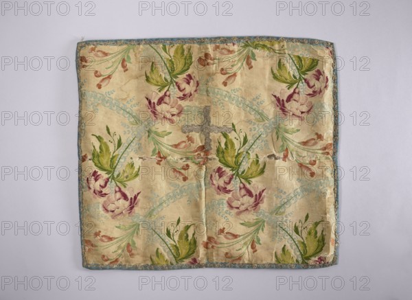 Velum;, church paraments; revindication provenance; flowers, leaves, plant ornaments; sacred art; churches and chapels furnishings; 18th century, France; liturgical parament; height 47 cm, width 54 cm, A square fabric panel with a pale beige background featuring a repeating floral pattern in shades of pink, burgundy, green, and light blue, edged with a narrow blue trim; the floral motifs include several large stylized blossoms in pink and burgundy with accompanying green leaves and small brownish-pink buds, arranged within curved light-blue decorative bands; the fabric shows visible creases, small tears, and areas of wear, and there is a small silver-gray decorative element near the center.