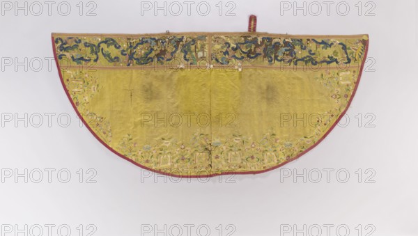 Church cope made of yellow silk fabric, embroidered along the edges with colorful floral sprigs around a winding ribbon; design of the motifs is difficult to read due to numerous patches. Pretext of green-yellow silk with an applied floral tendril motif. Liturgical vestment, 2nd half of the 18th century, Poland. Height 128 cm, width 280 cm., A semicircular textile displayed against a pale background, predominantly yellow with a narrow red border along the curved edge, showing a patterned top band with darker blue and green swirling motifs and scattered brown and gold areas, a main yellow field with subtle darker stains and creases, and a decorative lower edge featuring a row of floral embroidery in soft greens, pale blues, pinks, and cream tones, with a few small fabric ties along the upper edge and some visible wear and tiny holes near the center.