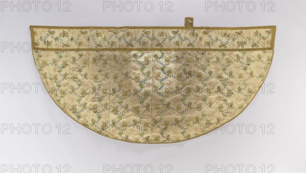 Church cope, creator unknown, origin unknown, liturgical vestment decorated with flowers, leaves and plant ornaments; used as church parament for churches and chapels; 18th century, France; height 142 cm, width 300 cm., A semicircular textile panel with a pale cream background patterned across its surface with a repeating floral and ribbon motif in colors including blue, green, pink, and gold, edged with a narrow gold trim and showing a horizontal band near the straight edge and a small rectangular tab at the top center, laid flat against a light gray background.