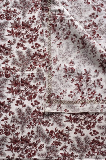 Pillowcase. Printed cotton canvas pillowcase with floral pattern on a white ground: printed sprigs arranged diagonally in smaller and larger bunches including roses, forget-me-nots, and bellflowers. Around the edges a printed winding snake with three dots in the bends. Colors: light brown and burgundy. 18th century, Province of Silesia 1742–1918. Height 84 cm, width 76 cm., The image shows a patterned fabric with a repeating floral design in red and white tones, featuring clusters of roses and leaves; a corner of the fabric is folded over, revealing the underside with a slightly faded version of the same floral motif and a decorative border in muted beige and red; the background is a light off-white and the overall texture of the cloth appears slightly worn with visible weave.