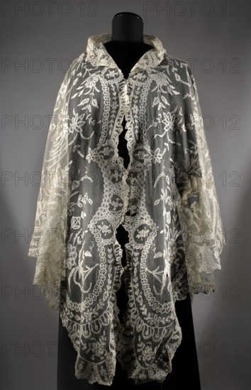 Cape, plant ornaments, floral ornaments, fashion, women's fashion, fashion accessories, capes, lace. Machine-made tulle background with a floral tendril pattern sewn from hand-made bobbin and partly needle-sewn lace. In the center is a motif of a small flower tree, surrounded by flower sprigs. 2nd half of the 19th century, Brussels. Clothing accessory, lace. Height 248 cm, width 123 cm., A sheer lace garment displayed on a black mannequin, featuring intricate floral and scrollwork embroidery in off-white and cream tones on a translucent net background, with a high collar and scalloped edges; the lace shows layered panels and flowing sleeves with delicate trim, and the backdrop is a neutral gray.