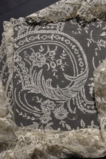 Cape, machine-made tulle background with floral tendril pattern sewn from hand-made, bobbin, and partly needle-sewn lace; center motif of a small flower tree surrounded by flower sprigs. Plant and floral ornaments; fashion, women's fashion, fashion accessory cape. 2nd half of the 19th century, Brussels. Lace; height 248 cm, width 123 cm., A piece of sheer embroidered fabric with intricate floral and foliate motifs displayed against a dark background; the fabric shows a large swirling floral pattern with stylized flowers, leaves, and vine-like tendrils, surrounded by scalloped and looped border embroidery, and edged by gathered lace ruffles; visible colors include off-white or cream embroidery and lace contrasted with a dark gray or black background.