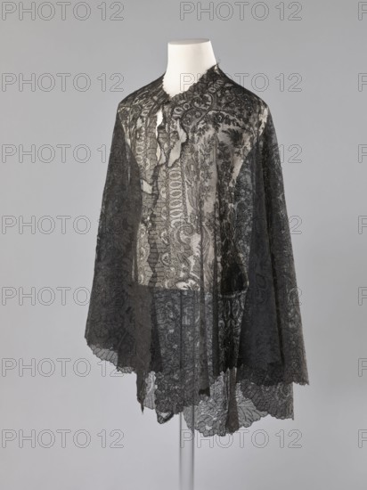 Cape - mantilla; unknown creator; shawl; purchase provenance; fashion; women's clothing; clothing accessory; lace mantilla cape, triangular. Pattern: garlands of leaves at the bottom; conical trees in the bends at the bottom. Late 19th century, France. Length 96 cm; width 344 cm., A mannequin torso draped with a sheer lace cape featuring intricate floral and scrollwork patterns, with scalloped edges and a high neckline; the lace is predominantly black layered over a lighter underlay that shows through, creating contrasts of black and grey against a neutral pale background.