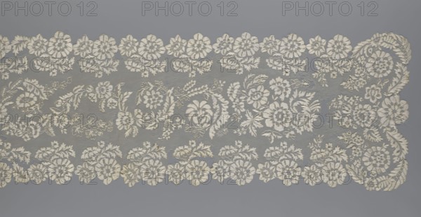 Shawl, shawl; clothing accessory; women's clothing; fashion; lace with serrated edges; design features: twigs with two flowers in the bends and curling flower twigs in the middle part; 19th century, Europe; length 48 cm, width 260 cm., The image shows a length of intricately patterned lace laid flat against a smooth background. The lace features a repeating floral motif with a scalloped edge composed of round flower shapes and leafy scrolls; between the scallops are flowing garlands of flowers and foliage woven into a fine net ground. A few larger floral clusters and stylized leaves form a central band, while several smaller blossoms and sprigs fill the spaces, creating an overall dense, delicate pattern. Visible colors include off-white for the lace and a cool gray for the background.
