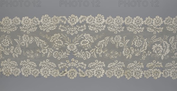 Shawl, shawl, clothing accessory, women's clothing, fashion, lace with serrated edges, design of twigs with two flowers in the bends and curling flower twigs in the middle part, 19th century, Europe, length 48 cm, width 260 cm., A horizontal strip of delicate lace with an intricate floral pattern in off-white and cream tones set against a muted gray background; the lace features a repeating arrangement of stylized flowers, leafy vines, and small blossoms forming a scalloped edge at top and bottom, with a few areas where the lace is slightly more densely stitched and several places showing subtle irregularities in the threads; overall the visible colors are off-white, cream, light beige, and gray.