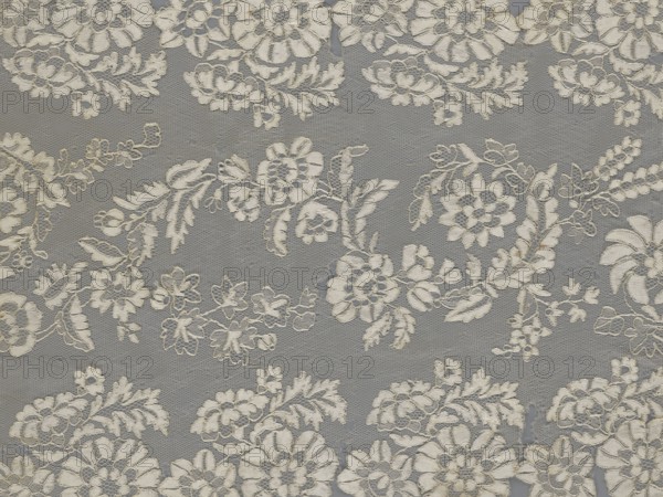 Shawl, shawl, clothing accessory, women's clothing, fashion, lace with serrated edges, design includes twigs with two flowers in the bends and curling flower twigs in the middle, 19th century, Europe, length 48 cm, width 260 cm., A close-up of a lace fabric showing a repeating floral pattern with embroidered flowers, leaves, and swirling vines on a sheer net background; the lace motifs appear in off-white and cream tones contrasted against a gray background, with visible texture from threads and occasional small gaps in the netting.
