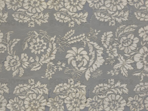 Shawl, shawl, clothing accessory, women's clothing, fashion, lace, serrated edges, in their bends twigs with two flowers, in the middle part curling flower twigs, 19th century, Europe, lace, length 48 cm, width 260 cm, A patterned textile with a repeating floral lace design in shades of off-white and beige set against a muted gray background, showing a group of embroidered flowers, leaves, and vine motifs interconnected across a fine netted mesh, with several circular and petal-shaped flower heads, feathery leaves, small clustered buds, and scalloped edges; the stitching creates subtle raised textures and shadows, and there are areas where the netting shows slight irregularities.