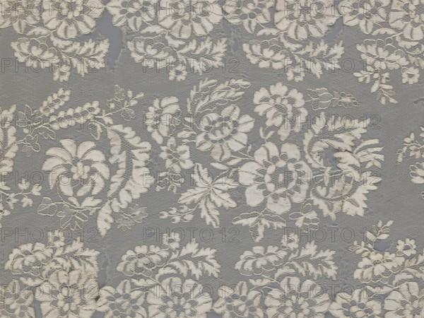 Shawl, shawl, clothing accessory, women's clothing, fashion, lace with serrated edges; design: twigs with two flowers in the bends and curling flower twigs in the central part; 19th century, Europe; length 48 cm, width 260 cm., A close-up view of lace fabric showing a repeating floral pattern with a group of embroidered flowers, leaves, and vines in off-white and cream tones set against a muted gray background, with areas where the open netting of the lace reveals the darker background and a few small irregular holes in the fabric.