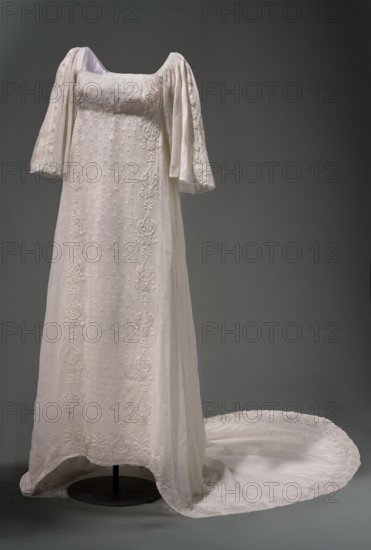 Dress with train, repossession provenance; Ludwika Queen of Prussia; 1776–1810 — souvenirs; trains clothing; geometric ornaments; Empire style; basket with flowers ornament. Dress of white muslin with a high waist and a long train, embroidered. Deep-cut square neckline, cinched at the front with a drawstring. Three-quarter set-in sleeves, gathered at the top and flared at the bottom. Skirt sewn to the bodice, narrow at the front and fuller at the back with a long train. Front decorated with embroidery: between two rows of stylized flower baskets are small scatterings of polka dots. Sleeves feature embroidered inserts with smaller geometric patterns. Hem and train decorated with stylized flower baskets. Early 19th century, Silesia. Women's clothing; length 131 cm front, length 230 cm back, circumference 408 cm hem., A long white dress displayed on a stand against a neutral gray background, made of sheer lightweight fabric with a flowing train pooling on the ground; the gown has a square...