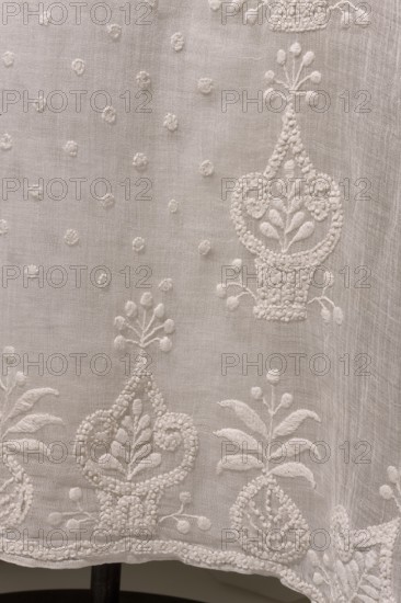 Dress with train, Dress of white muslin, early 19th century, Silesia. High waist, with a long train; deep-cut square neckline, cinched at the front with a drawstring. Three-quarter set-in sleeves, gathered at the top and flared at the bottom, with embroidered inserts featuring smaller geometric patterns. Skirt sewn to the bodice, narrow at the front and fuller at the back, long train. Front decorated with embroidery: between two rows of stylized flower baskets are small scatterings of polka dots. Hem and train decorated with stylized flower baskets. Empire style. Trains clothing, geometric ornaments, basket with flowers ornament. Length 131 cm front, 230 cm back; hem circumference 408 cm. Associated with Ludwika Queen of Prussia; 1776–1810 — souvenirs; repossession, A close-up view of a textured light-colored fabric with embroidered floral and botanical motifs in the same pale color, showing several ornate vase-like shapes, leaves, small round raised dots, and delicate stitched borders...